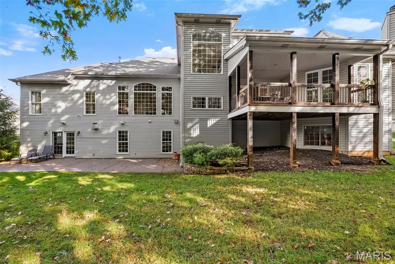 Rear elevation featuring multi-level outdoor living spaces including a covered upper deck with wooden posts and a paved ground-level patio, expansive lawn, siding exterior, and multi-pane windows with arched detailing — 112 Castlewood Estates Drive, O'Fallon, MO 63368