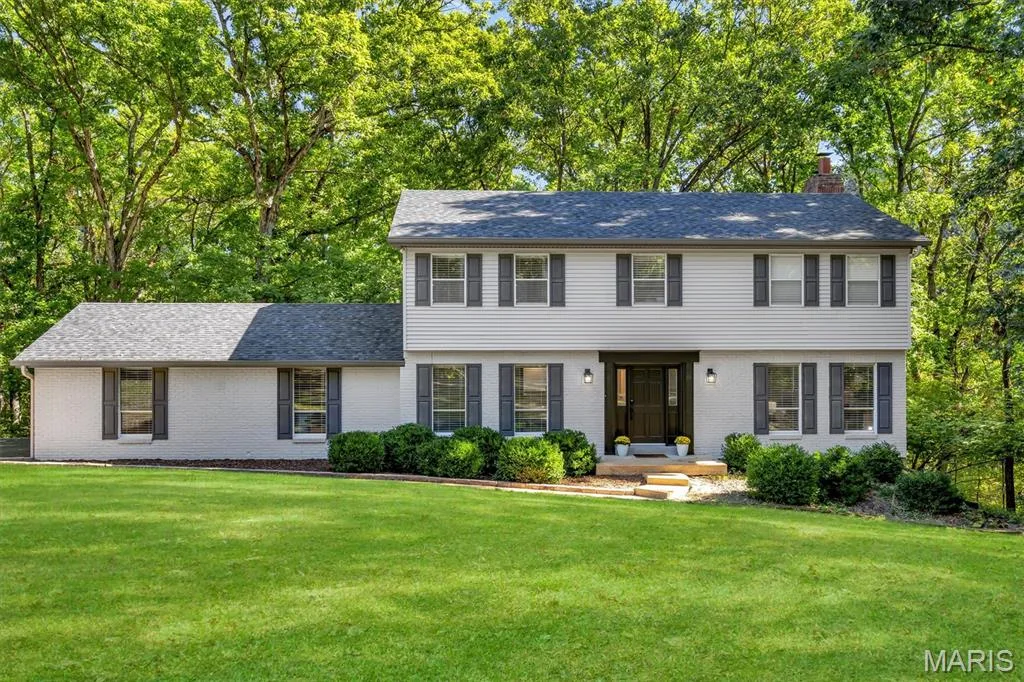 Two-story exterior featuring light-toned brick and horizontal siding, dark-framed multi-pane windows, decorative shutters, and a shingled roof, set on a manicured lawn with mature tree canopy — 1444 Eagle Ridge Road, Wildwood, MO 63038