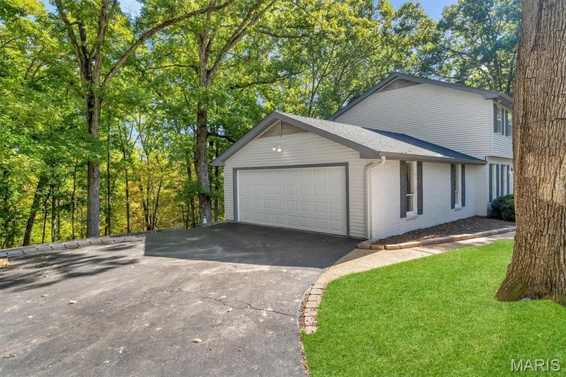 Exterior featuring a multi-level structure with light-toned siding and brick accents, an attached two-car garage, asphalt driveway, and mature surrounding woodland — 1444 Eagle Ridge Road, Wildwood, MO 63038