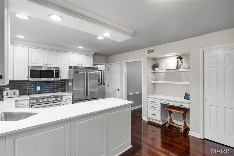 Kitchen featuring white cabinetry, solid-surface countertops, a dark tile backsplash, stainless steel appliances, and a built-in workstation with integrated shelving — 1444 Eagle Ridge Road, Wildwood, MO 63038