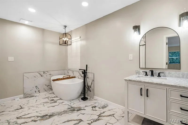 Freestanding soaking tub with matte black floor-mounted faucet, vanity with stone countertop, arch-top mirror, and large-format patterned stone-finish flooring — 1444 Eagle Ridge Road, Wildwood, MO 63038