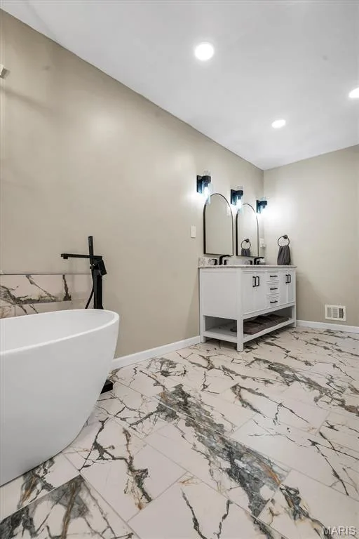 Freestanding soaking tub with a matte black floor-mounted faucet, vanity with a neutral countertop and dual undermount sinks, arched wall mirrors, and large-format tile flooring with veining detail — 1444 Eagle Ridge Road, Wildwood, MO 63038