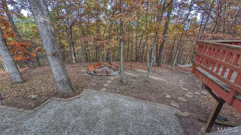 Wooded backyard featuring a gravel patio, stone paver pathway, built-in circular fire pit area with wooden benches, and an elevated wood deck with railing — 1444 Eagle Ridge Road, Wildwood, MO 63038