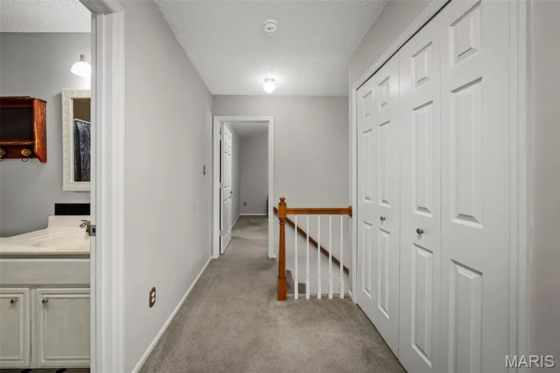 Carpeted hallway with painted walls, white bi-fold closet doors, and wood newel post with white balusters — 445 Lantana Lane, St Peters, MO 63376