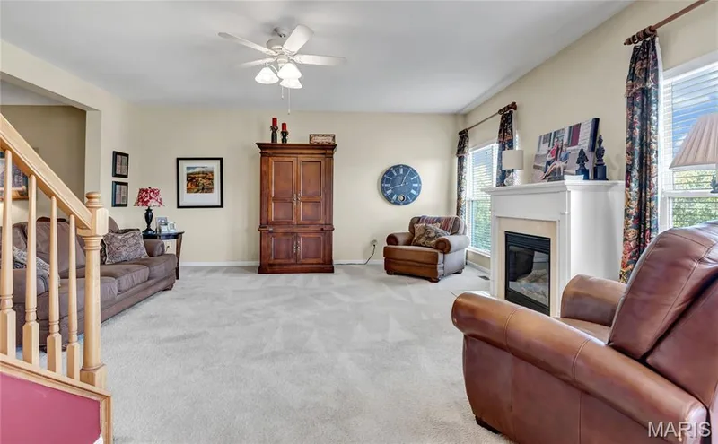 Living area featuring a fireplace with a white mantel, neutral wall-to-wall carpeting, a ceiling fan with integrated lighting, and wooden staircase railing — 2061 Avalon Mist Circle, Dardenne Prairie, MO 63368