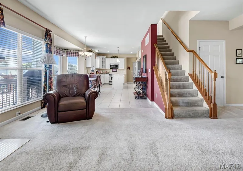 Carpeted living area featuring large windows, staircase with wooden handrails and balusters, and tile flooring in the kitchen area with white cabinetry — 2061 Avalon Mist Circle, Dardenne Prairie, MO 63368