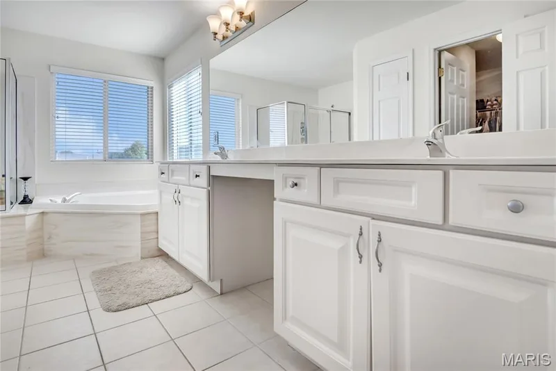 Bathroom featuring a dual-sink vanity with white cabinetry, a large wall-mounted mirror, ceramic tile flooring, and a jetted soaking tub with a tile surround — 2061 Avalon Mist Circle, Dardenne Prairie, MO 63368