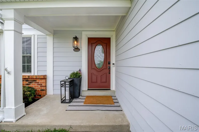 Covered concrete porch featuring a red entry door with an oval glass insert, a black wall-mounted lantern light fixture, white siding, and a brick foundation — 1094 Harness Drive, St Charles, MO 63304