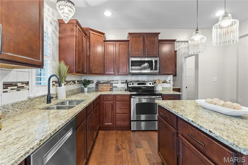 Kitchen featuring wood cabinetry, stone-finish countertops, stainless steel appliances, wood-finish flooring, and tiered glass pendant lighting — 3222 Starkville Street, St Charles, MO 63301