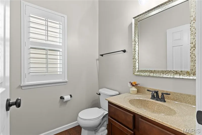 Bathroom featuring a vanity with wood-finish cabinetry, a solid-surface countertop with an integrated sink, a window with plantation shutters, wood-finish flooring, and a framed mirror — 3222 Starkville Street, St Charles, MO 63301