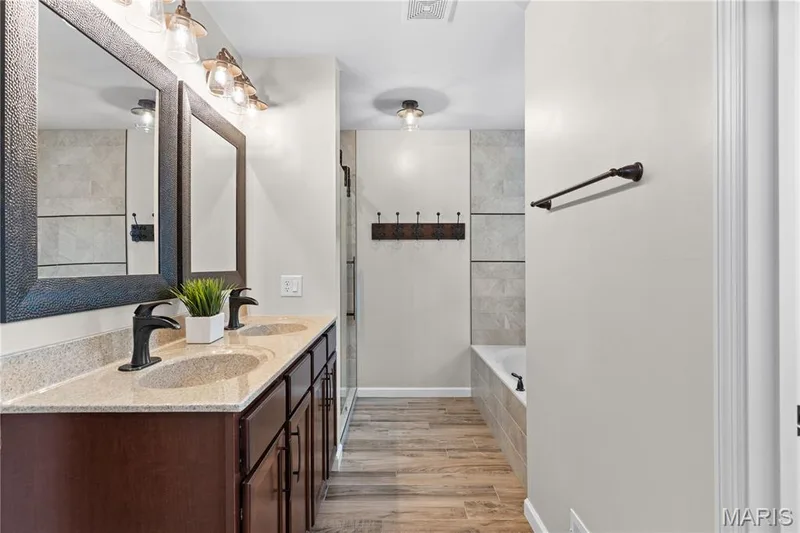 Double vanity featuring a light-toned countertop, dual under-mount sinks, wood-finish cabinetry, dark-finished hardware, and wood-finish flooring — 3222 Starkville Street, St Charles, MO 63301