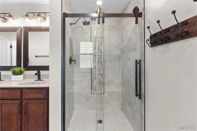 Dual-sink vanity with stone countertop and dark wood cabinetry, glass-enclosed shower with floor-to-ceiling tile, vertical mosaic accent inlay, and dark matte hardware throughout — 3222 Starkville Street, St Charles, MO 63301
