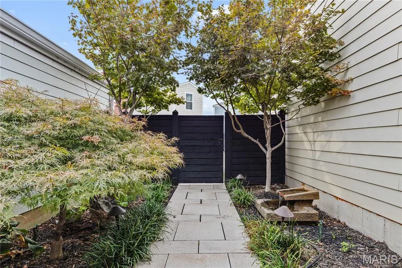 Landscaped side yard featuring a stone paver walkway, black horizontal privacy fencing, mature ornamental trees, and a tiered stone water feature — 3222 Starkville Street, St Charles, MO 63301