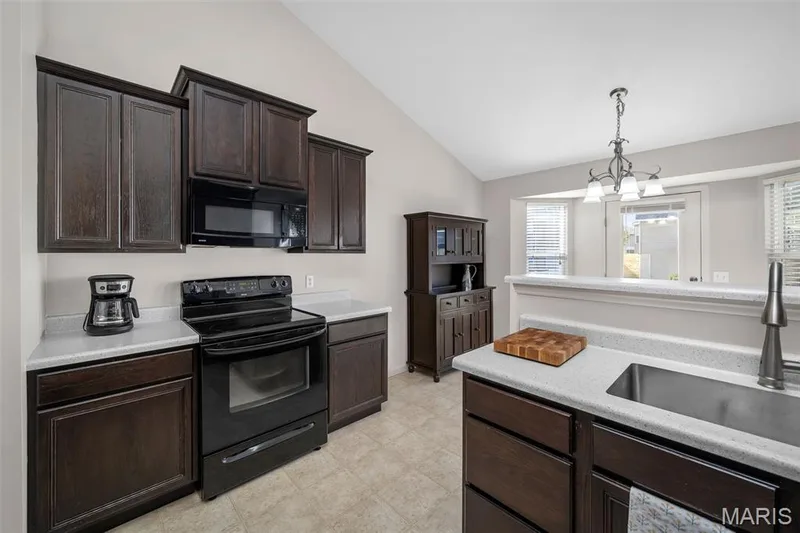 Kitchen featuring dark stained cabinetry, light-toned solid surface countertops, black appliance suite, vaulted ceilings, and a stainless steel undermount sink with a high-arc pull-down faucet — 757 Lost Canyon Boulevard, Wentzville, MO 63385