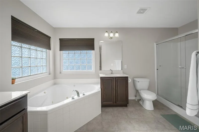 Ensuite featuring a tiled soaking tub, separate glass-enclosed shower, wood-finish vanity with a stone countertop, and textured glass windows — 757 Lost Canyon Boulevard, Wentzville, MO 63385