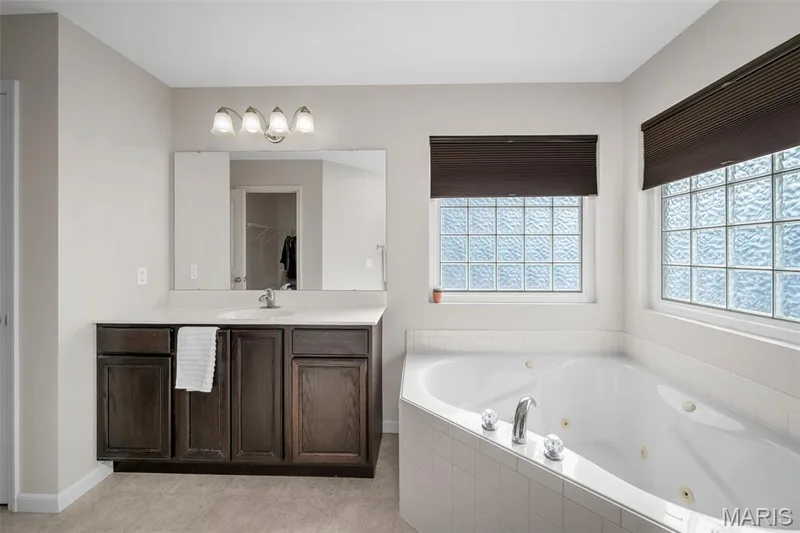 Master Bath featuring a dark-toned wood vanity with a light-colored countertop, an integrated jetted soaking tub finished with white tile, a large wall-mounted mirror, four-light vanity fixture, and windows with privacy glass — 757 Lost Canyon Boulevard, Wentzville, MO 63385