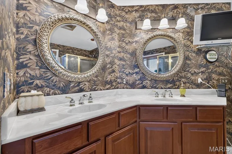 Dual-sink vanity with a solid surface countertop, warm-toned wood cabinetry, dual ornate circular wall mirrors, and multi-light fixtures — 69 Weule Boat Harbor, St Charles, MO 63301