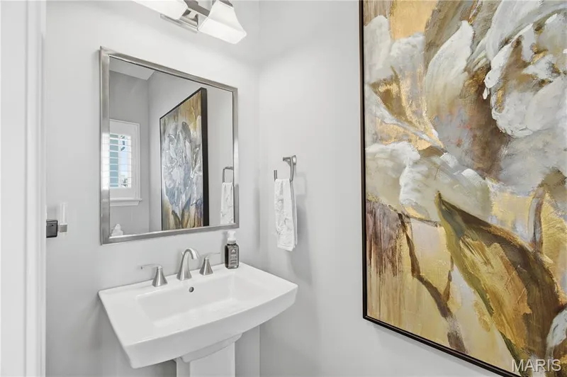 Wall-mounted vanity mirror with a brushed metal frame, white pedestal sink, dual-handle metal faucet, and two-light wall sconce fixture — 17028 Fire Glow Drive, Wildwood, MO 63011