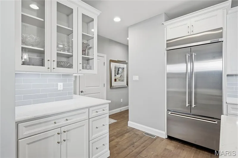 White cabinetry with glass-front upper displays, stone-finish countertops, subway tile backsplash, built-in stainless steel refrigerator, and wood-finish flooring — 17028 Fire Glow Drive, Wildwood, MO 63011