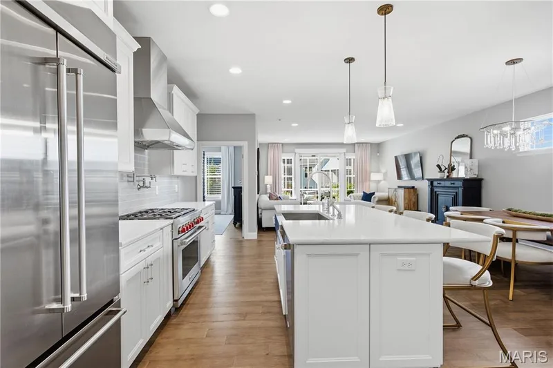 Kitchen featuring white shaker-style cabinetry, quartz countertops, a central island with an integrated sink, stainless steel professional-style range with wall-mounted pot filler, and wood-finish flooring — 17028 Fire Glow Drive, Wildwood, MO 63011