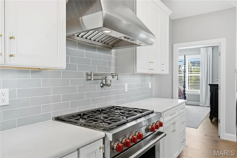 White shaker-style cabinetry featuring gold-tone hardware, stainless steel range with a professional-grade hood, light-colored stone-finish countertops, subway tile backsplash, and wall-mounted pot filler — 17028 Fire Glow Drive, Wildwood, MO 63011