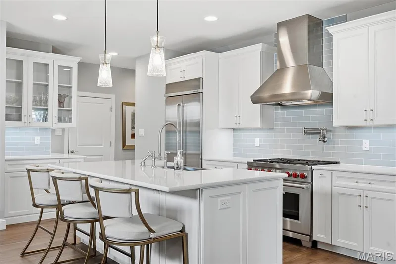 Kitchen featuring white cabinetry, an expansive central island with stone-finish countertop, stainless steel appliances including a professional-grade range and chimney-style vent hood, subway tile backsplash, and two glass pendant lights — 17028 Fire Glow Drive, Wildwood, MO 63011
