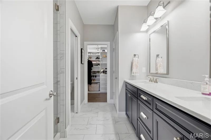 Bathroom featuring a dual-sink vanity with dark cabinetry and stone-finish countertops, tile flooring, walk-in tiled shower, and built-in closet shelving — 17028 Fire Glow Drive, Wildwood, MO 63011