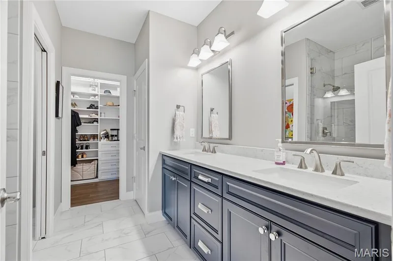 Double vanity featuring navy blue cabinetry, light-toned stone-finish countertops, undermount sinks, and polished chrome hardware, complemented by floor-to-ceiling built-in closet shelving and tile flooring — 17028 Fire Glow Drive, Wildwood, MO 63011