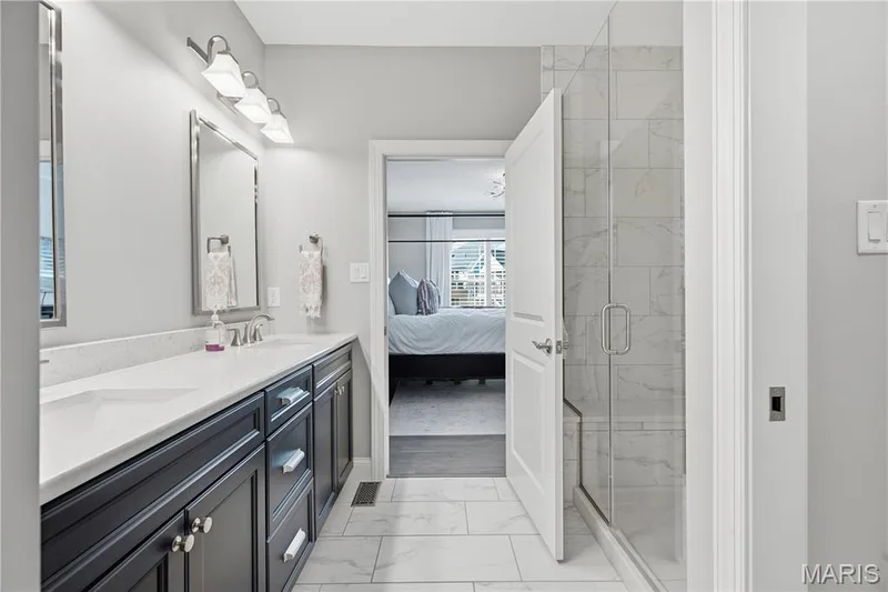 Bathroom featuring a dark-toned dual vanity with a light-colored solid surface countertop, a walk-in shower with a glass enclosure and floor-to-ceiling tile, and rectangular tile flooring — 17028 Fire Glow Drive, Wildwood, MO 63011