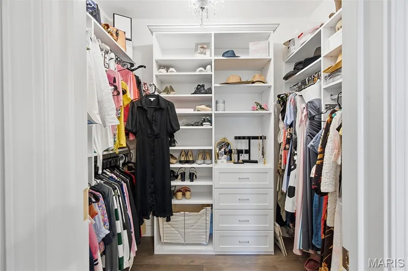 Walk-in closet featuring custom floor-to-ceiling white shelving, integrated multi-drawer cabinetry, overhead crystal-style light fixture, and wood-finish flooring — 17028 Fire Glow Drive, Wildwood, MO 63011